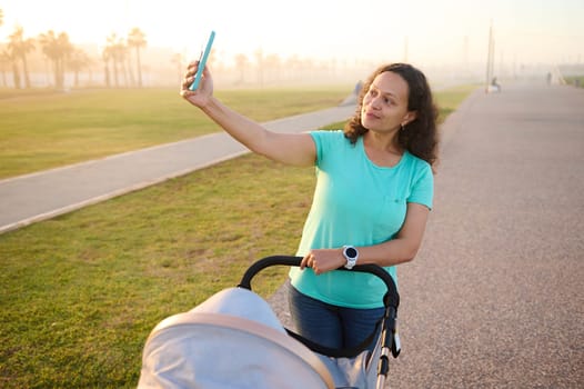 Smiling young woman recording video, taking selfie while pushing pram with her baby, strolling on the promenade at sunset. Happy mother enjoying walk with her child on a summer day. Maternity leave