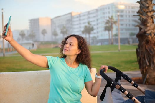 Beautiful woman taking selfie on her mobile phone while pushing pram with her sleeping newborn baby, on the city promenade at sunset. Happy young mother enjoying walk with her child on a summer day