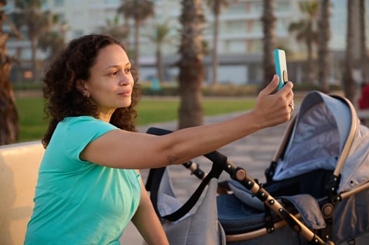 Young curly haired woman 40s, mother sitting on city bench while walking with her baby in pram, taking a self portrait on her smartphone, sharing her maternity leave lifestyle on social media content