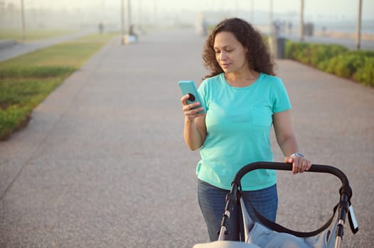 Beautiful young woman 40s, happy mother using mobile phone, checking application or online shopping, browsing websites, watching webinars while pushing baby pram on the promenade at sunset.