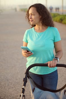 Vertical shot of a curly multi ethnic happy young woman 40s, loving mother pushing baby pram, smiling looking aside, standing on the footpath on the promenade at sunset with a smartphone in her hands