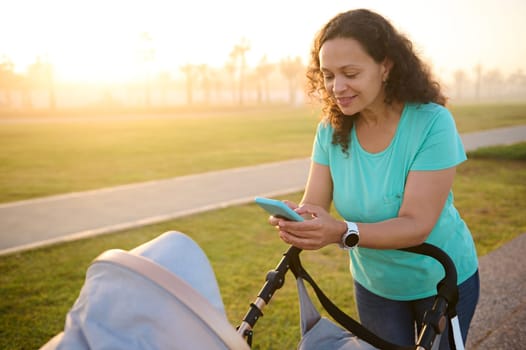 Young pretty woman, mother using mobile phone while pushing baby pram with her baby, standing outdoors at sunset, Happy mom shopping online, taking photo of her newborn child sleeping in baby buggy