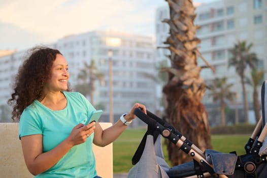 Smiling happy mother holding using a smart mobile phone, sitting on city bench and pushing baby pram on the promenade at sunset. People. Maternity leave lifestyle. Online communication