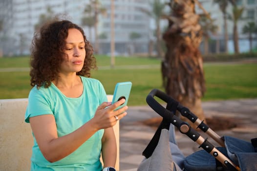 Woman using mobile phone outdoors, sitting on a bench near a baby pram with sleeping baby. People. Maternity. Online communication