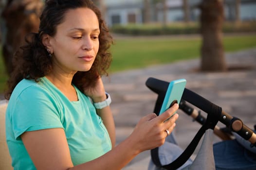 Confident portrait of a beautiful woman, young mother sitting near a baby pram with her sleeping child, using smart mobile phone while walking at sunset. People. Maternity leave lifestyle concept.