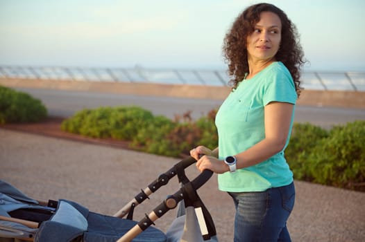 Young mother dreamily looking aside, walking with her baby sleeping in pram, standing on the footpath on the promenade against Atlantic ocean on the background at sunset. Maternity leave lifestyle