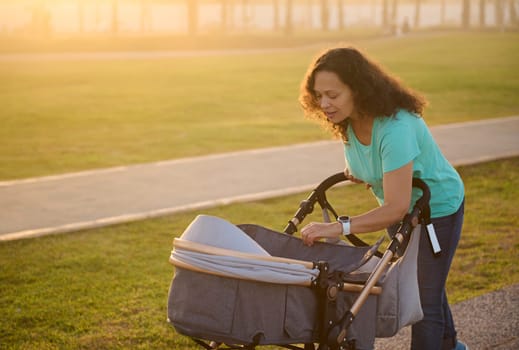 Smiling happy young multi ethnic woman, loving caring mother walking with her newborn baby in stroller, spending time funny on the city park t sunset