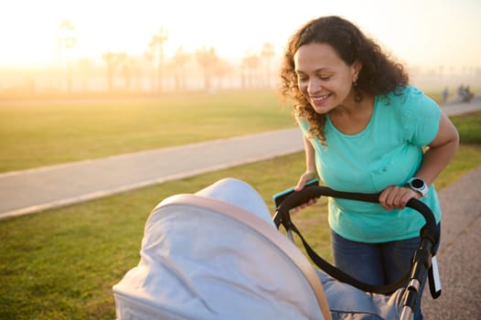 Cheerful and happy young woman 40s, loving caring mother looking into baby carriage while walking in the park at sunset, dressed in blue turquoise t-shirt ang denim. Sunbeam of sunset on background