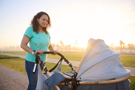 Beautiful young mother pushing stroller with her baby, looking at him while he's sleeping in the pram during daily walking outdoors at beautiful sunset time. Cheerful motherhood, care, parenthood.