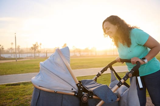 Side portrait of authentic happy mother looking and pushing infant baby stroller while walking in park at sunset. The concept of healthy active lifestyle. Daily outdoors walking and maternity leave.