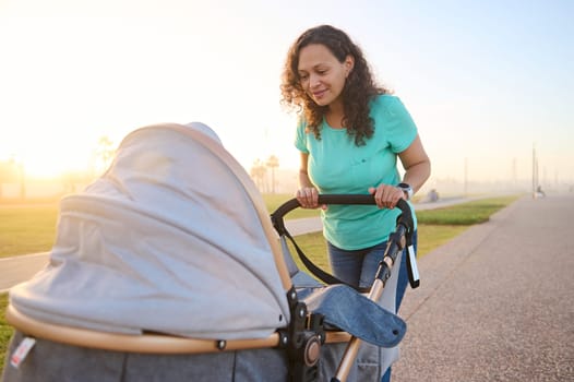 Smiling beautiful and young woman mother and baby in a stroller walking in the park at sunset background. Infancy and maternity leave lifestyle