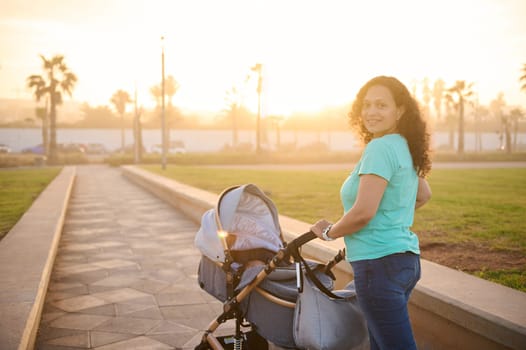 Side portrait happy smiling young mother looks at camera, pushes silver gray baby carriage. Infant sleeping and breathing fresh air in summer at sunset. Family. Motherhood. Maternity leave lifestyle