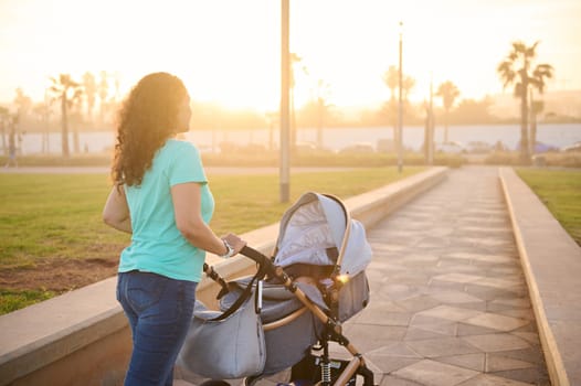Rear portrait of young mother pushing stroller with her sleeping lovely little baby, walking in the promenade at sunset. Beautiful sunbeams on the background. Motherhood, healthy lifestyle concept.