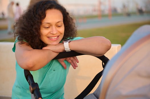 Portrait of happy mother smiling broadly to her baby in the baby pram. Multi ethnic curly haired brunette woman in blue turquoise t-shirt walking with her newborn child sleeping in the baby carriage