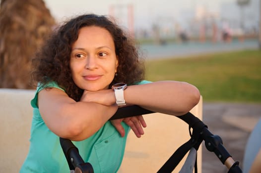 Portrait of Middle Eastern beautiful woman, young mother smiling looking aside, putting her hands on the baby pram with her sleeping child, walking at sunset. People. Maternity leave lifestyle concept