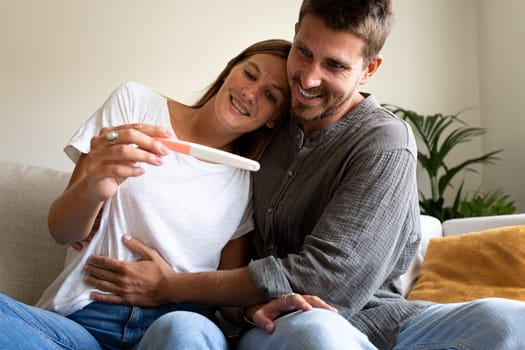 Young happy caucasian couple looking at positive pregnancy test together at home living room. Husband touching wife belly.Pregnancy and family concept.