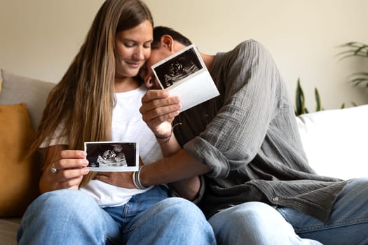 Happy and excited pregnant couple looking baby ultrasounds at home sitting on the couch. Man touching wife belly. Selective focus. Love and family concept.