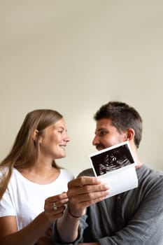 Vertical portrait of pregnant couple looking at each other in love while holding baby ultrasounds. Selective focus on hand. Love and family concepts.