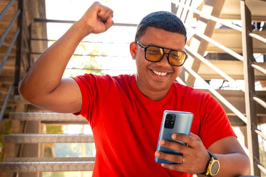 Latin man wearing excited sunglasses using mobile phone watching a football match. Student elated by the result of the exam.