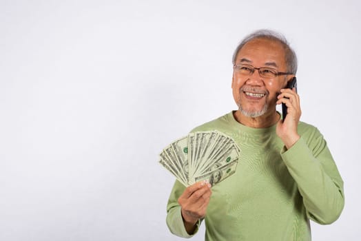 Portrait Asian smiling old man is holding a bunch of money fan and talking on cell mobile phone studio shot isolated on white background. happy elderly hold dollar banknotes