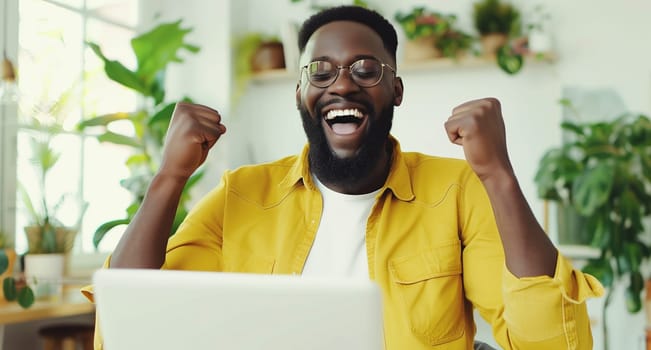 Happy overjoyed laughing african man celebrating success raising his hands up while working on laptop sitting at home office