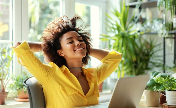 Happy relaxed young african woman enjoying sitting at desk during working at the laptop at home with green plants, female freelancer or student resting