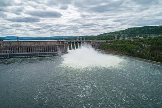 Aerial view of concrete dam releasing water into river on cloudy day. Water discharge at hydroelectric power plant.