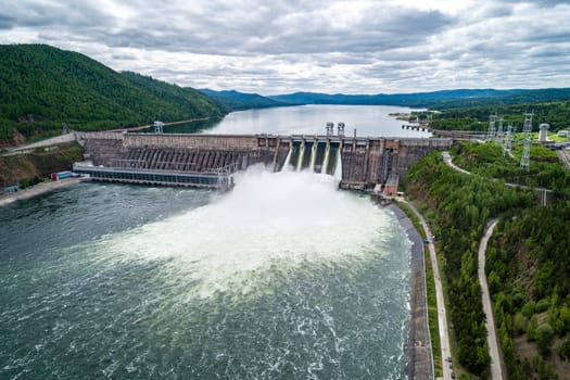 Aerial view of concrete dam releasing water into river on cloudy day. Water discharge at hydroelectric power plant.