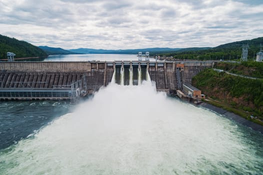 Aerial view of concrete dam releasing water into river on cloudy day. Water discharge at hydroelectric power plant.