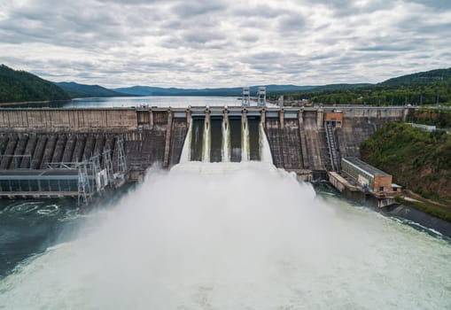 Aerial view of concrete dam releasing water into river on cloudy day. Water discharge at hydroelectric power plant.