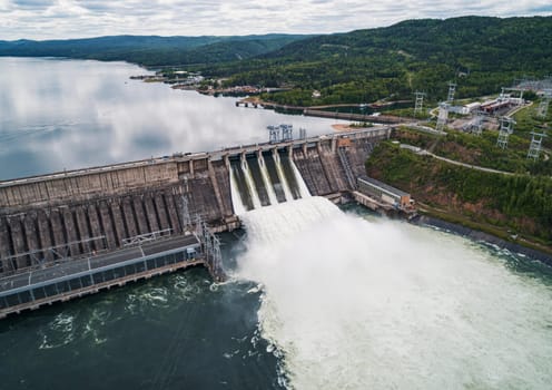 Aerial view of concrete dam releasing water into river on cloudy day. Water discharge at hydroelectric power plant.