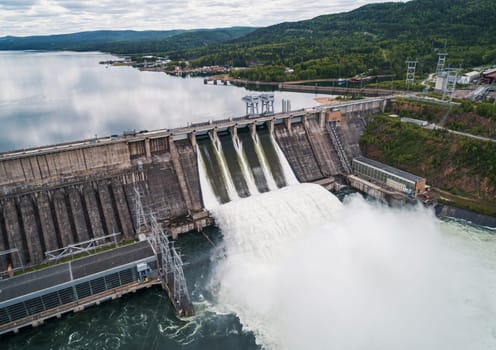 Aerial view of concrete dam releasing water into river on cloudy day. Water discharge at hydroelectric power plant.