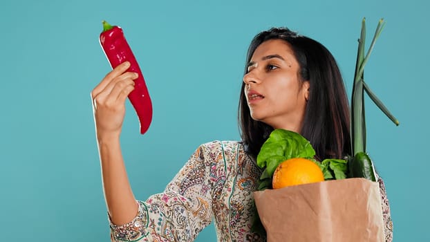 Indian woman with ecological paper bag in hands looking at bell pepper, living healthy lifestyle. Ecology lover holding shopping bag with organic produce, studio backdrop, camera B