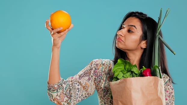 Indian woman with paper bag with vegetables and fruits testing quality, looking at orange, studio background. Vegetarian verifying groceries are ripe after buying them from zero waste shop, camera B