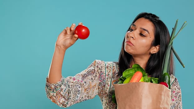 Woman with recycled paper bag in hands filled with vegetables testing quality, studio background. Sustainable living person inspecting groceries after doing purchases in shop, camera B