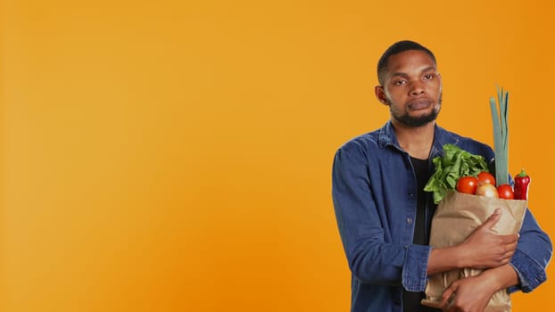 Portrait of african american guy holding a paper bag full of bio food, buying organic locally grown fruits and vegetables. Confident vegan person supporting healthy eating and zero waste. Camera B.