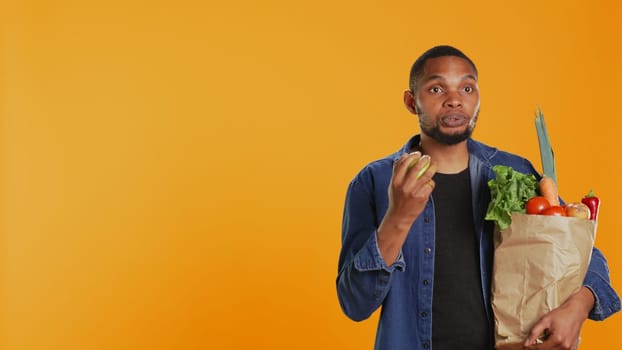 Confident man juggling with an eco friendly green apple on camera, presenting ripe locally grown fruits. Young adult playing around and carrying paper bag with organic produce. Camera B.