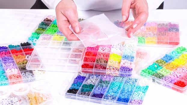 Woman’s hands gracefully poised over a collection of beads, sorted by color in transparent organizers. The array of beads spans a vibrant spectrum, from deep purples to bright oranges, meticulously arranged for easy selection as she embarks on creating a custom piece of jewelry.