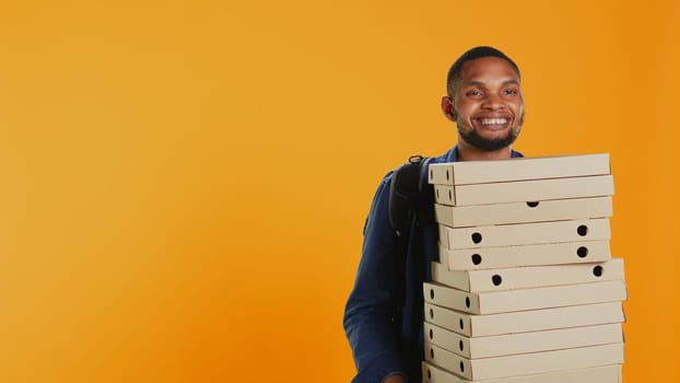 Male pizzeria courier carrying huge pile of pizza boxes in studio, preparing to deliver meal order to customers. Young deliveryman holding a big stack of fast food takeaway, shipping. Camera B.