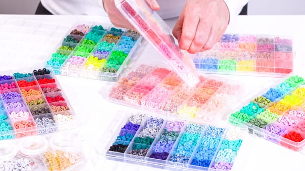 Woman’s hands gracefully poised over a collection of beads, sorted by color in transparent organizers. The array of beads spans a vibrant spectrum, from deep purples to bright oranges, meticulously arranged for easy selection as she embarks on creating a custom piece of jewelry.