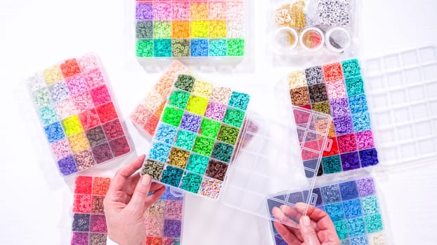 Flat lay. Woman’s hands gracefully poised over a collection of beads, sorted by color in transparent organizers. The array of beads spans a vibrant spectrum, from deep purples to bright oranges, meticulously arranged for easy selection as she embarks on creating a custom piece of jewelry.