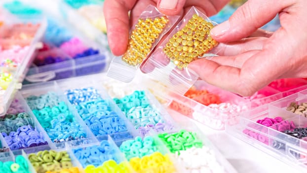 Woman’s hands gracefully poised over a collection of beads, sorted by color in transparent organizers. The array of beads spans a vibrant spectrum, from deep purples to bright oranges, meticulously arranged for easy selection as she embarks on creating a custom piece of jewelry.