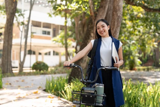 Smiling Asian businesswoman holding a laptop and standing with her bicycle outdoors, promoting eco-friendly commuting and professional success