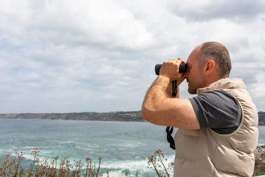 Mockup Mature Man, Adult Looks Into Distance Through Binoculars At Sea, Ocean Shore. Blue Sky, Water On Background. Summer Vacation, Hobby. Handsome 40 Yo Caucasian Male Explores Nature During Trip.