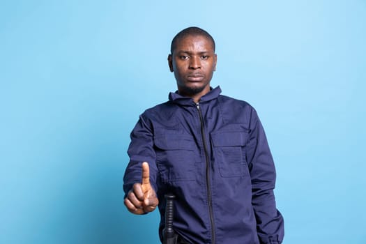 African american security guard poses with a finger up in studio, presenting a restriction sign against blue background. Person working as a patrol officer showing a stop symbol in studio.