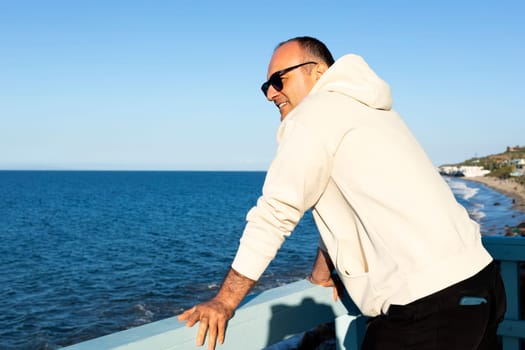 Happy Middle Age Caucasian Man Standing Alone On Wooden Pier And Staring At Sea, Ocean At Sunny Day. 40 Yo Male Wears Hoodie, Denim, Sunglasses. Vacation, Trip, Relaxation. Peaceful Atmosphere