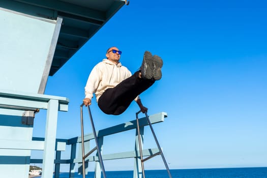 Middle Age Caucasian Man Doing Physical Exercise Pulling Legs Up At Beach Of Sea, Ocean At Sunny Day, Blue Sky On Background. Adult In Hoodie, Denim, Sunglasses Does Sport In Unusual Unequipped Place.