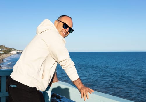 Smiling Middle Age Caucasian Male Stands Alone On Wooden Pier. Blue Sea, Ocean, Sky on Background. Sunny Day. Man Wears Hooded Sweatshirt, Jeans, Sunglasses. Vacation Relaxation. Peaceful Atmosphere