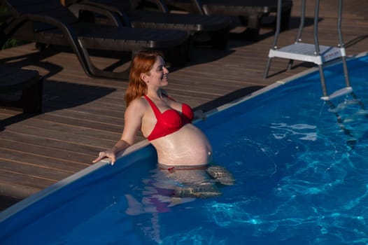 A red-haired pregnant woman in a red bikini is resting in the pool, leaning on the side