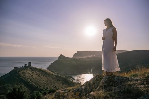 A woman stands on a rocky hill overlooking the ocean. She is wearing a white dress and she is enjoying the view. The scene is serene and peaceful, with the sun shining brightly in the background